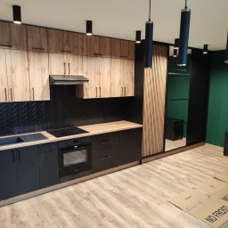 Kamil Wąsik - Nowoczesna kuchnia z drewnianymi górnymi szafkami, czarnymi dolnymi szafkami i czarną ścianą z płytek ułożonych w jodełkę, obok szafa z lustrzanymi drzwiami i drewnianymi lamelami, podłoga wyłożona...