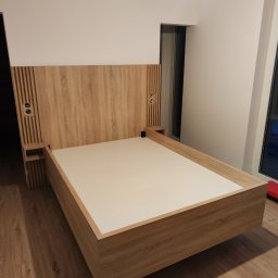 Kamil Wąsik - Łóżko z drewnianym wezgłowiem z wbudowanymi szafkami nocnymi i gniazdkami, w minimalistycznym wnętrzu z drewnianą podłogą.