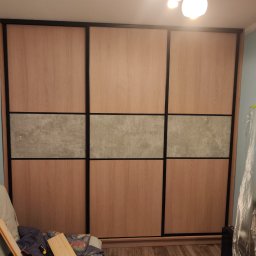 Kamil Wąsik - Trzydrzwiowa szafa wnękowa z przesuwnymi drzwiami, jasne drewno i szary panel dekoracyjny, widoczne narzędzia i elementy montażowe na pierwszym planie.