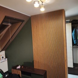 Kamil Wąsik - Drewniana ścianka działowa z pionowych listew oddzielająca część sypialni od garderoby, widoczna szafa z ubraniami, skośny sufit i lampa sufitowa.