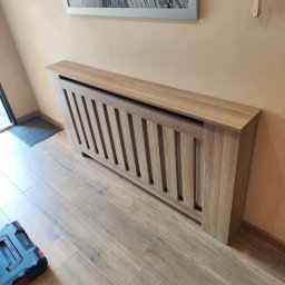 Kamil Wąsik - Drewniana obudowa grzejnika z pionowymi szczeblami, dopasowana do podłogi z paneli, widoczna skrzynka narzędziowa w rogu.