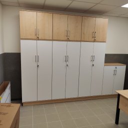 Kamil Wąsik - Wysokie szafy zamykane na klucz, białe dolne moduły i drewnopodobne górne, ustawione w rzędzie wzdłuż ściany, widoczne z boku biurko i karton z boku.