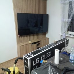 Kamil Wąsik - Montaż telewizora OLED na ścianie z drewnianą boazerią i szafką RTV, widoczny karton po telewizorze i odkurzacz, wnętrze w trakcie urządzania.