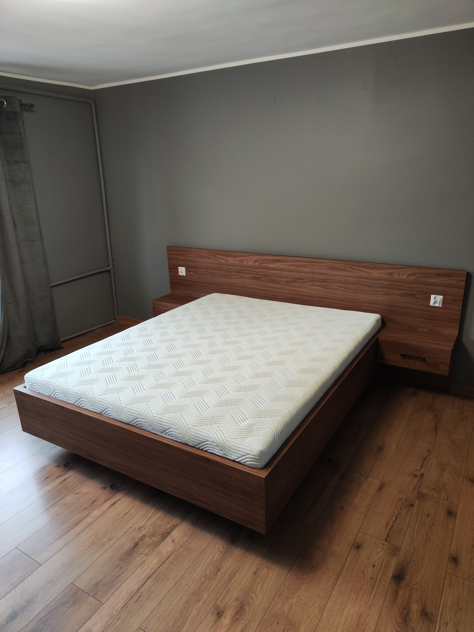 Nowoczesne łóżko z drewna orzechowego z wbudowanymi szafkami nocnymi i gniazdkami elektrycznymi, materac z delikatnym wzorem, na podłodze panele imitujące drewno.