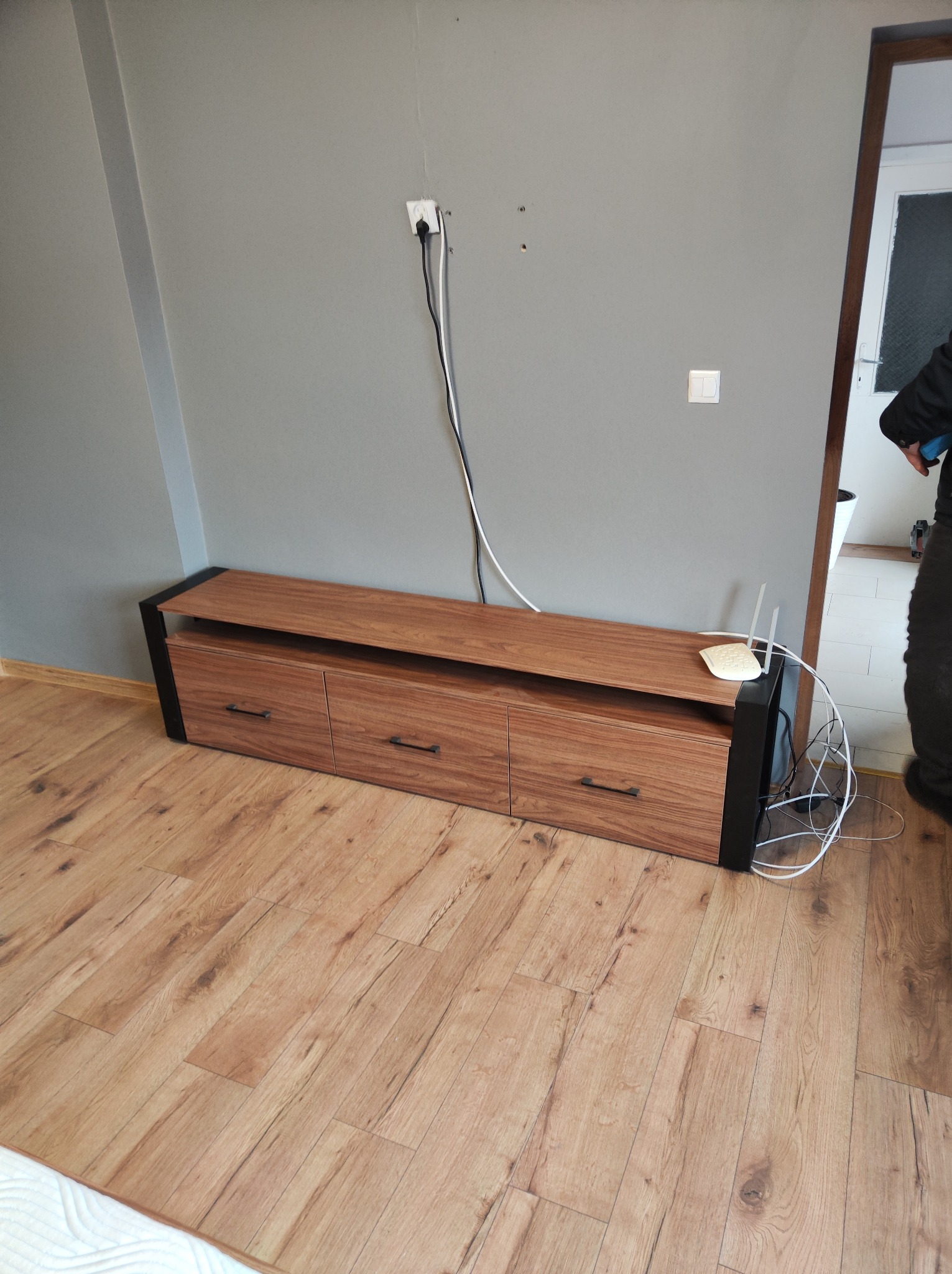 Brązowa szafka RTV z trzema szufladami i czarnymi metalowymi nogami, router Wi-Fi na blacie, szara ściana z wystającymi kablami i gniazdkiem elektrycznym