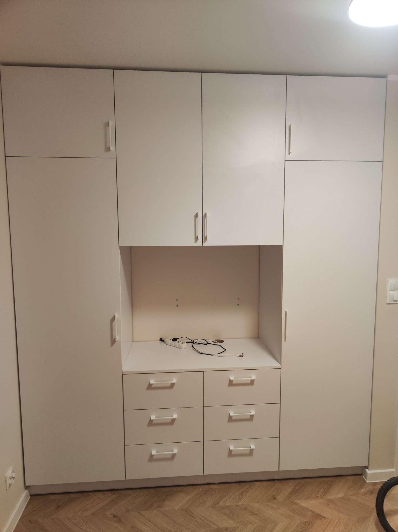 Biała zabudowa meblowa z szafkami górnymi i dolnymi z szufladami, z otwartą przestrzenią na blacie z widocznymi kablami, na podłodze parkiet ułożony w jodełkę.