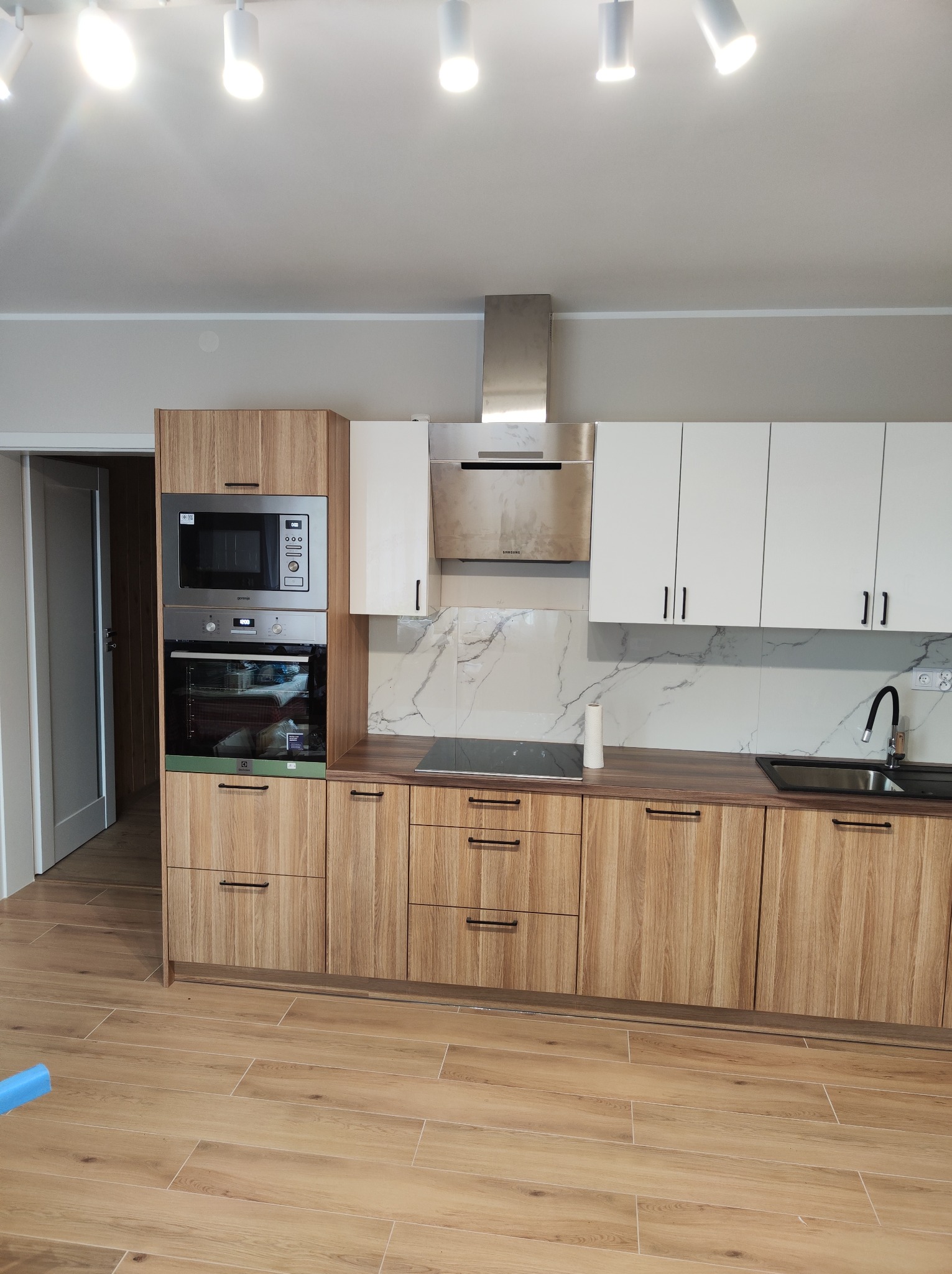 Nowoczesna kuchnia z frontami w kolorze drewna i białymi szafkami górnymi, czarne uchwyty, marmurowy backsplash, sprzęt AGD zabudowany w wysokiej szafce, płyta indukcyjna i czarny zlew.