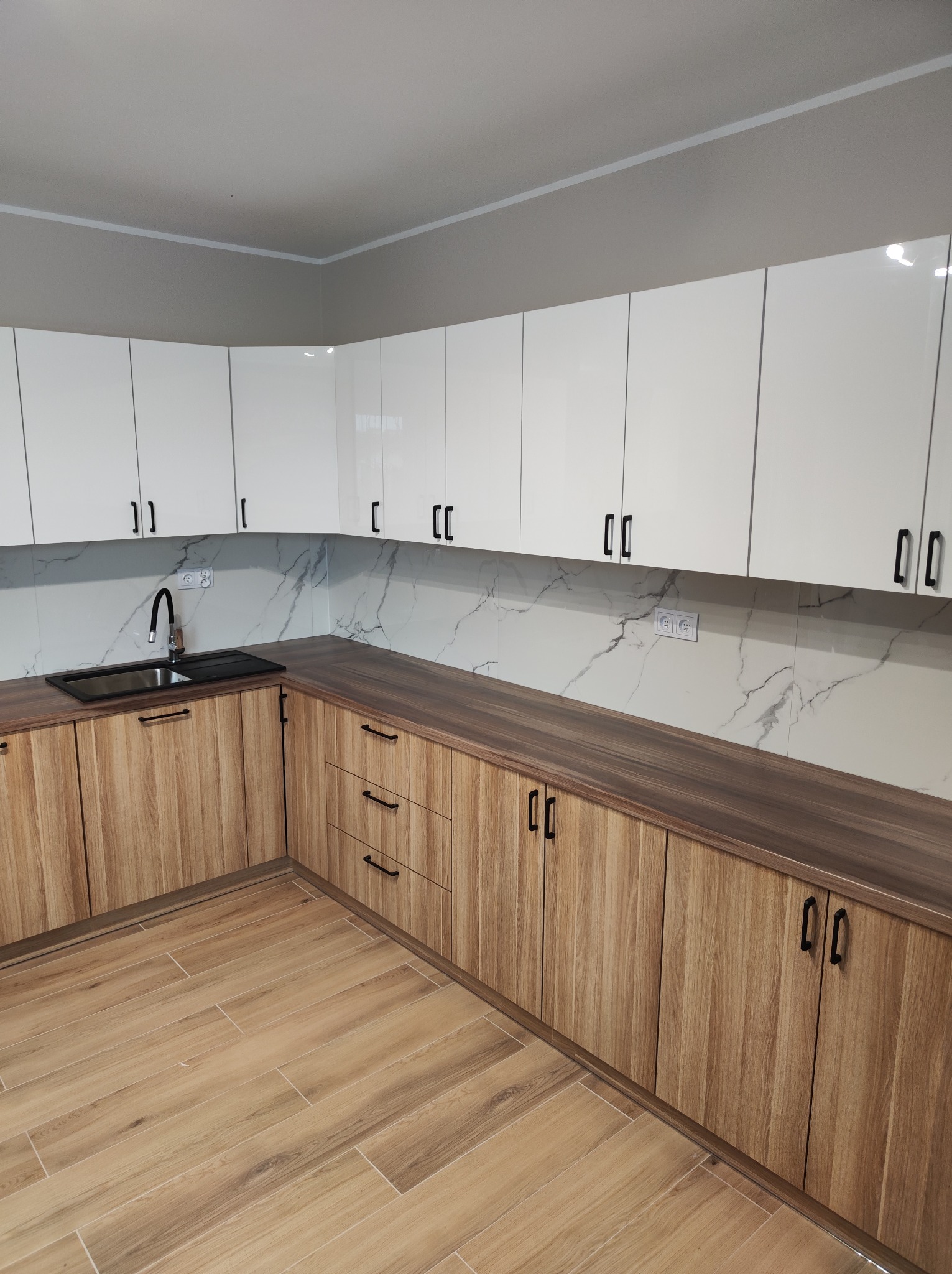 Nowoczesna kuchnia z frontami w kolorze jasnego drewna i białymi szafkami górnymi z czarnymi uchwytami; blat w ciemnym odcieniu brązu, ściana nad blatem wykończona płytkami imitującymi marmur.