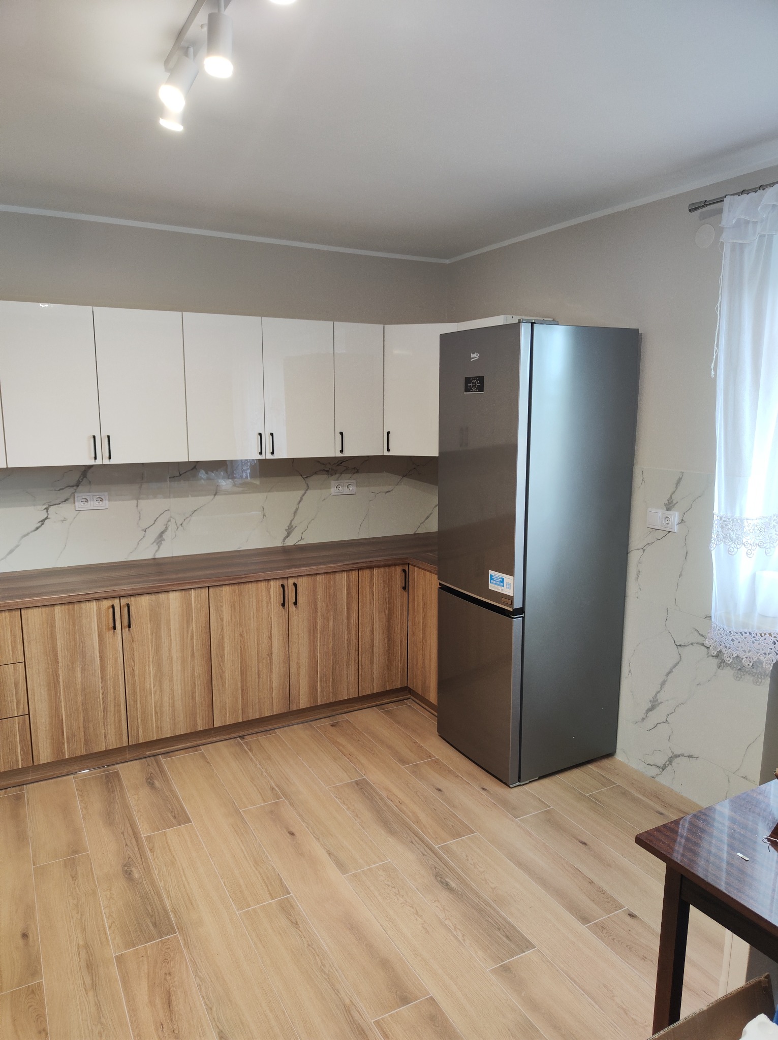 Jasna kuchnia z drewnianymi dolnymi szafkami i białymi górnymi, marmurowy backsplash, srebrna lodówka Beko, drewniana podłoga i firanka w oknie.