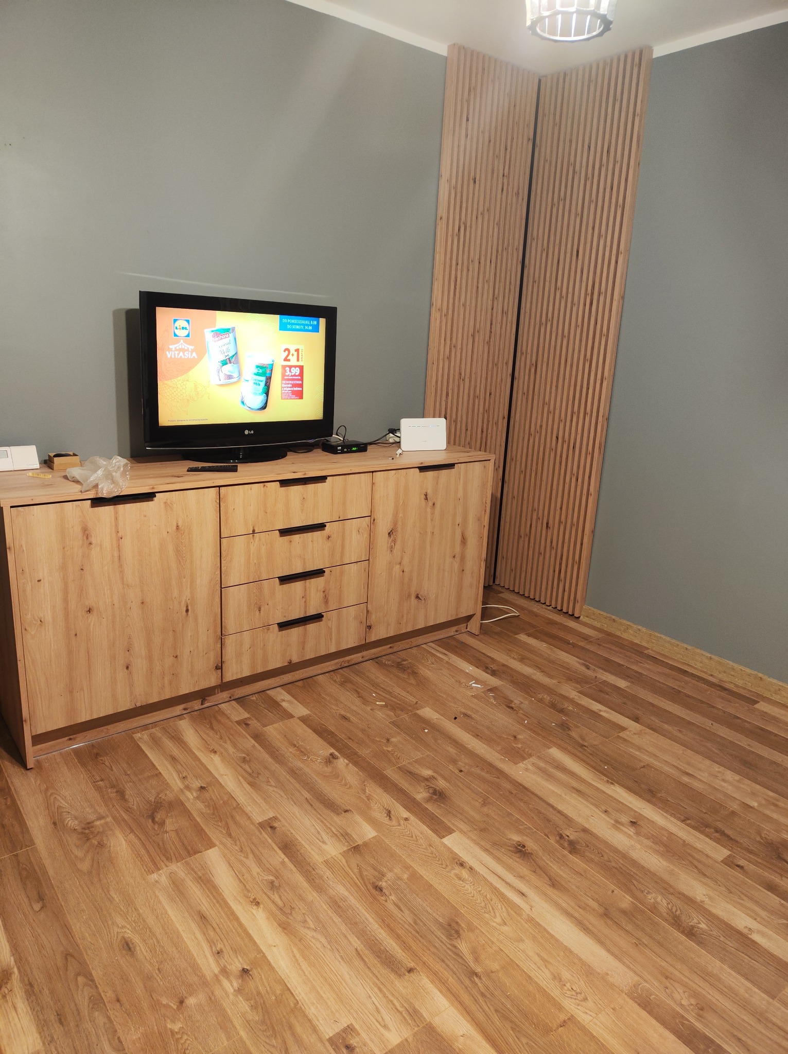 Drewniana komoda RTV z czarnymi uchwytami na tle szarej ściany i drewnianych paneli ściennych. Na komodzie stoi telewizor z włączoną reklamą.