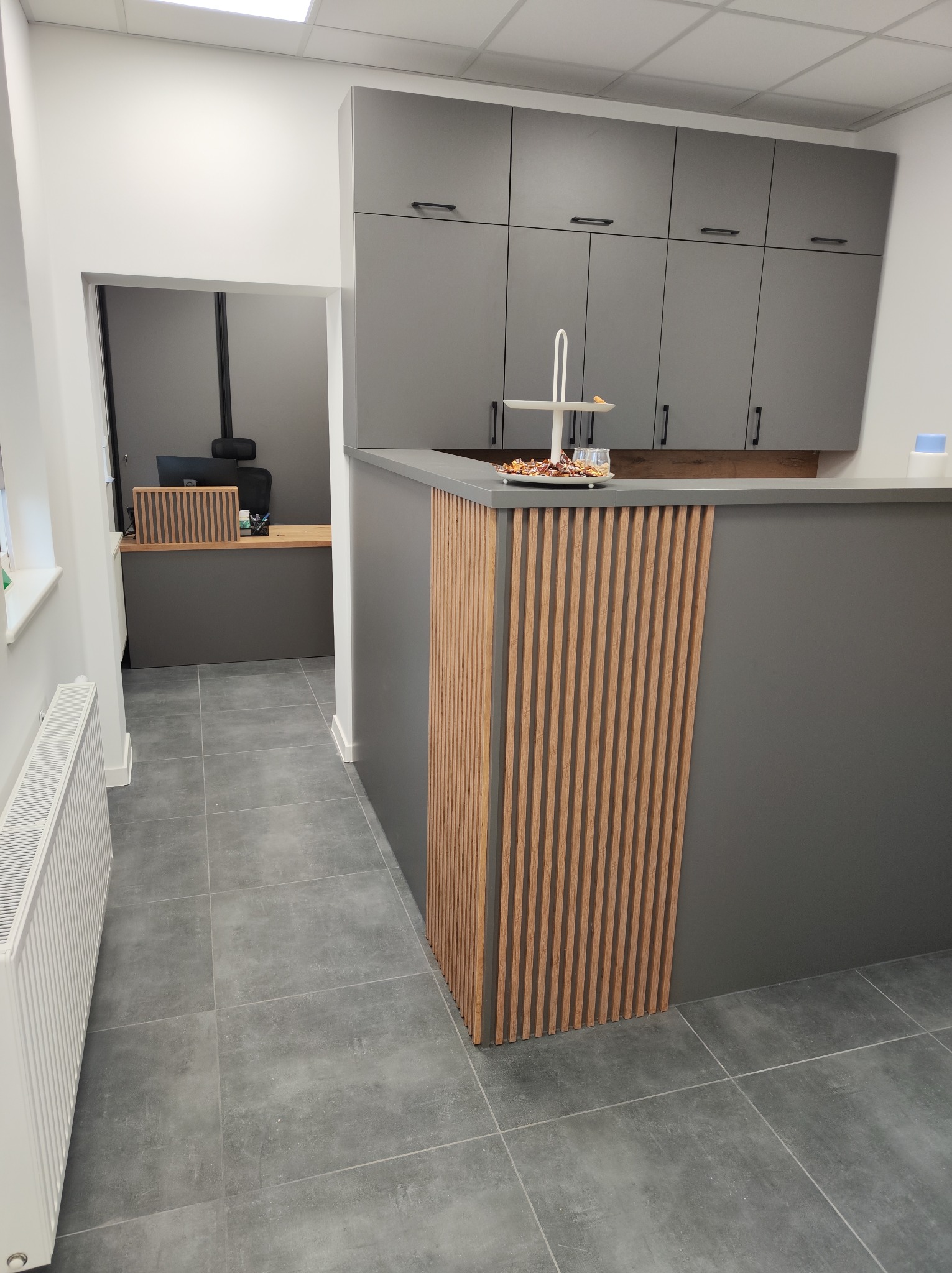 Szara zabudowa kuchenna z szafkami wiszącymi i blatem, na którym stoi etażerka z przekąskami. Front baru wykończony pionowymi drewnianymi listwami. W tle widoczne biuro z drewnianym biurkiem.