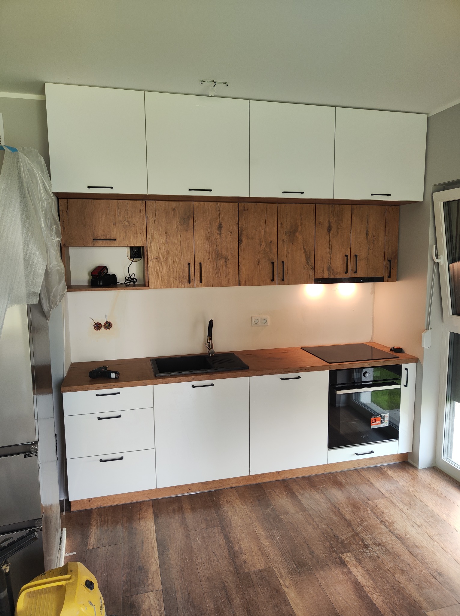 Nowoczesna kuchnia na wymiar w Chlebowie z białymi i drewnopodobnymi szafkami, czarnym zlewem i płytą indukcyjną, widoczne narzędzia na blacie i odkurzacz w rogu, sugerujące trwający montaż.