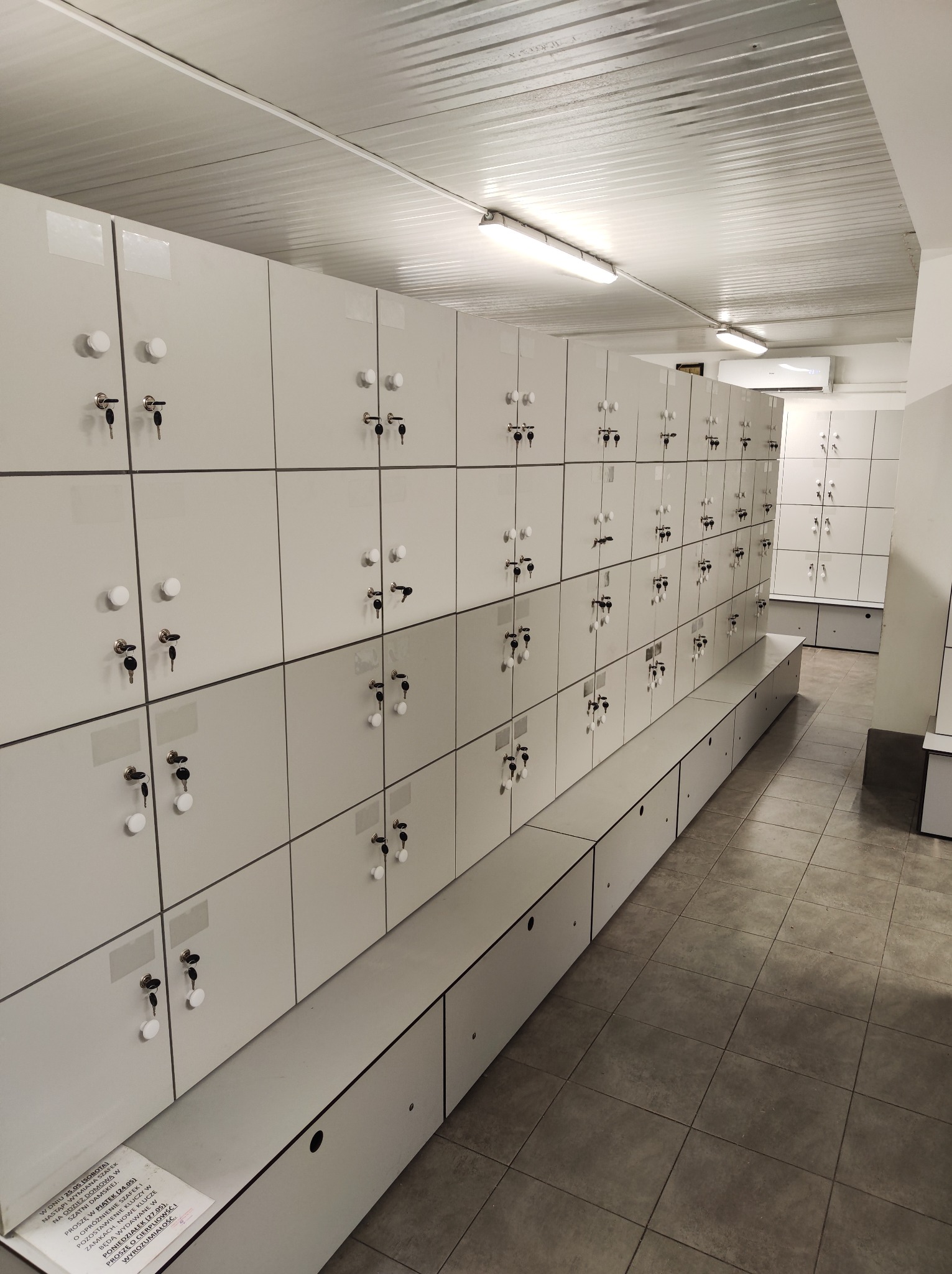 Rząd białych szafek zamykanych na klucz w szatni, widoczne klucze w zamkach, na podłodze płytki imitujące kamień, na jednej z szafek kartka z informacją o wymianie szafek.