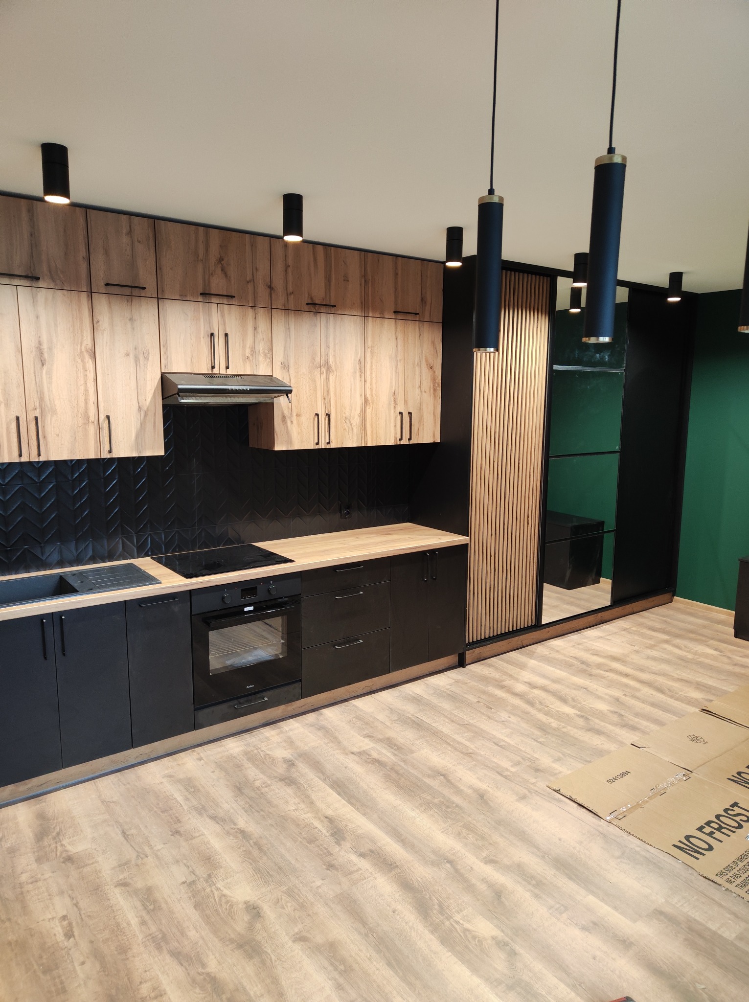 Nowoczesna kuchnia z drewnianymi górnymi szafkami, czarnymi dolnymi szafkami i czarną ścianą z płytek ułożonych w jodełkę, obok szafa z lustrzanymi drzwiami i drewnianymi lamelami, podłoga wyłożona...