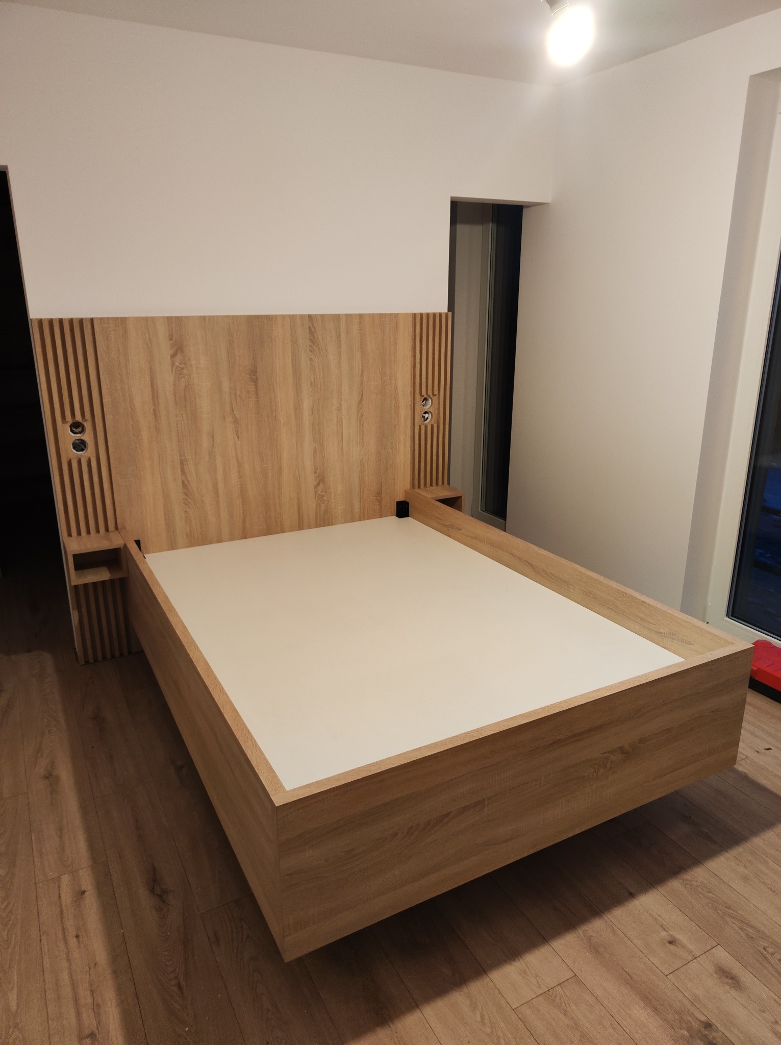 Łóżko z drewnianym wezgłowiem z wbudowanymi szafkami nocnymi i gniazdkami, w minimalistycznym wnętrzu z drewnianą podłogą.