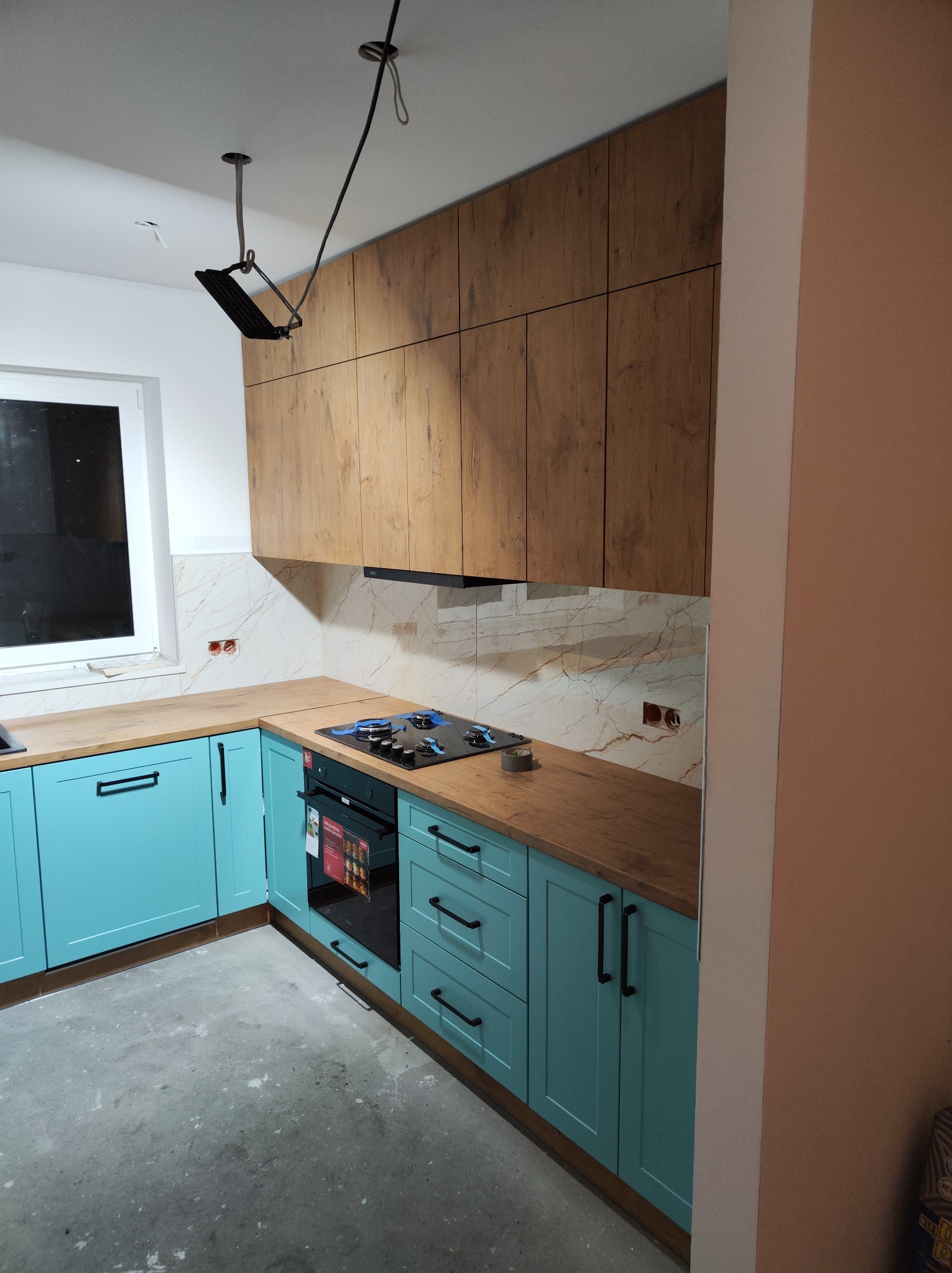Niebieska zabudowa kuchenna z drewnianym blatem i górnymi szafkami z imitacją drewna, płyta indukcyjna zamontowana, widoczne kable od oświetlenia pod sufitem, surowa podłoga przed położeniem paneli.
