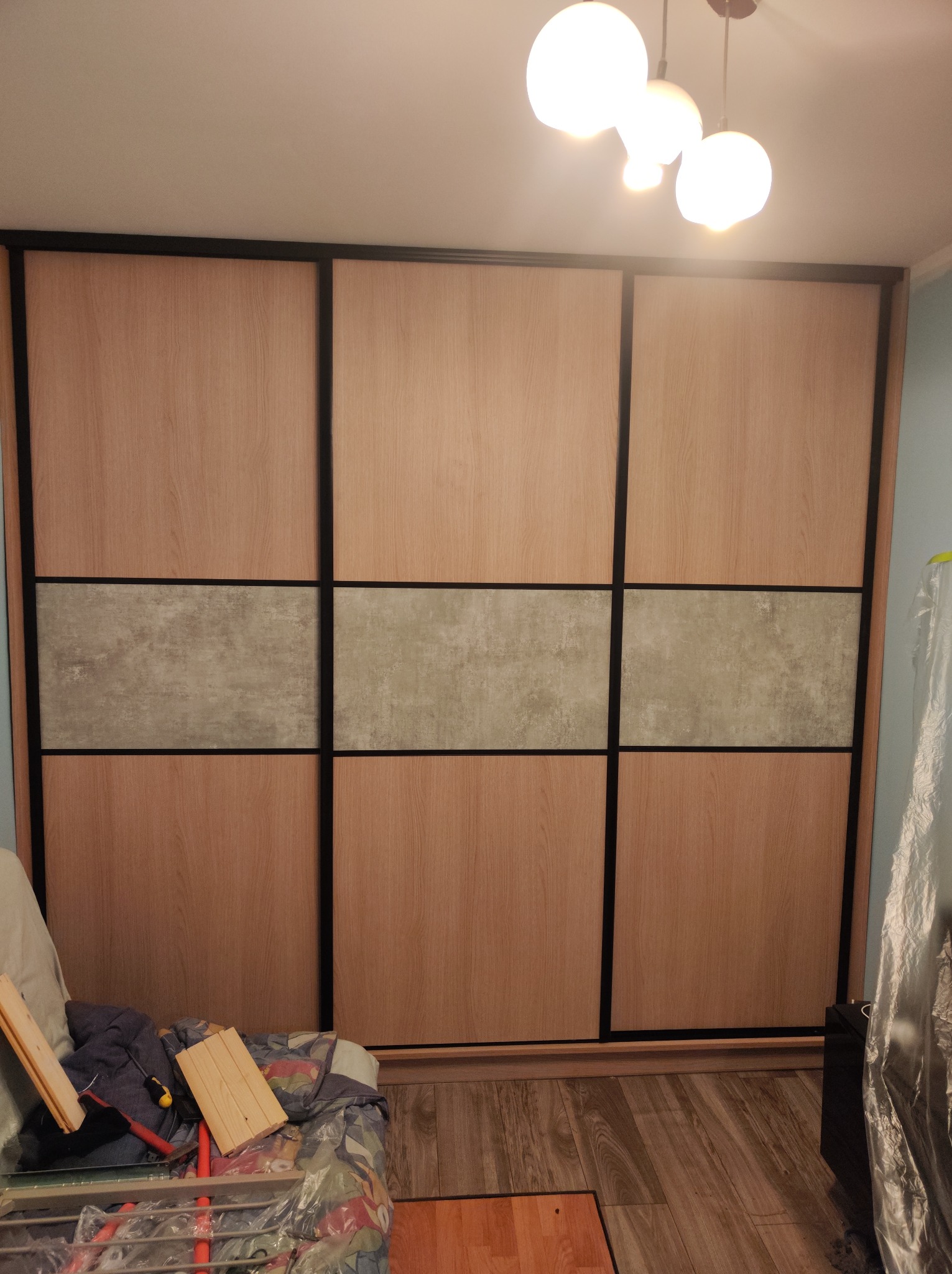 Trzydrzwiowa szafa wnękowa z przesuwnymi drzwiami, jasne drewno i szary panel dekoracyjny, widoczne narzędzia i elementy montażowe na pierwszym planie.