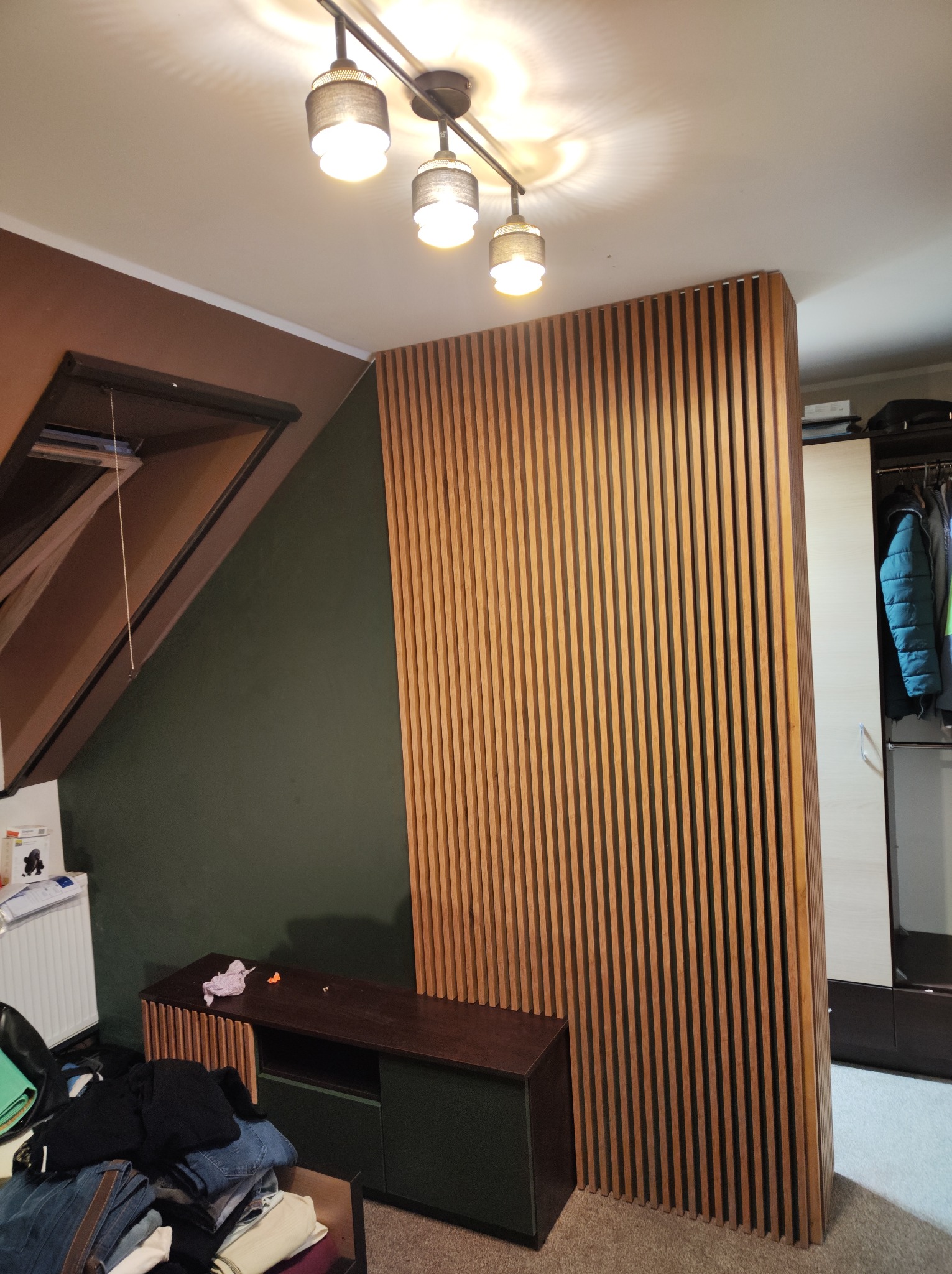 Drewniana ścianka działowa z pionowych listew oddzielająca część sypialni od garderoby, widoczna szafa z ubraniami, skośny sufit i lampa sufitowa.