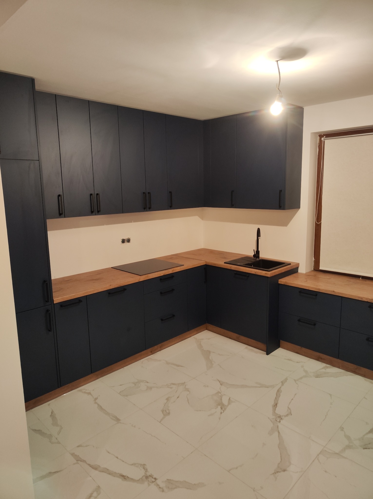 Nowoczesna kuchnia z ciemnymi, granatowymi szafkami i drewnianym blatem, czarny zlew i bateria, marmurowe płytki podłogowe, surowa żarówka zwisająca z sufitu.