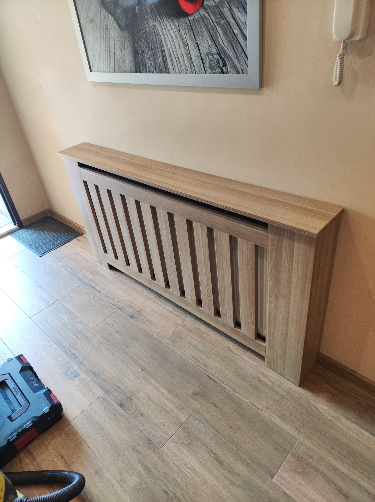 Drewniana obudowa grzejnika z pionowymi szczeblami, dopasowana do podłogi z paneli, widoczna skrzynka narzędziowa w rogu.