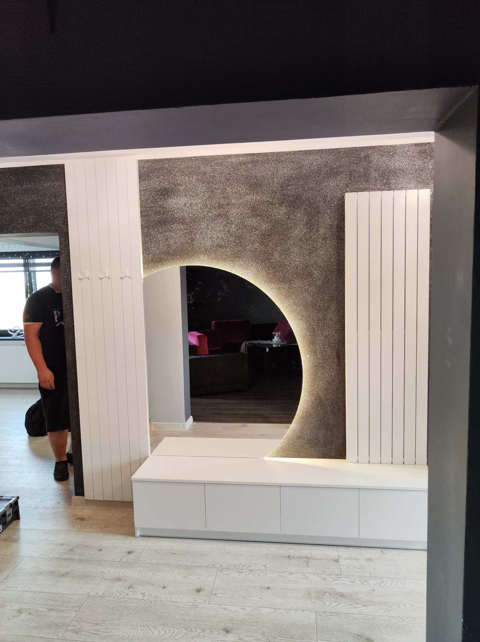 Nowoczesny przedpokój z lustrem w kształcie półksiężyca z podświetleniem LED, białą szafką i panelem z wieszakami; ściana z efektem brokatu.