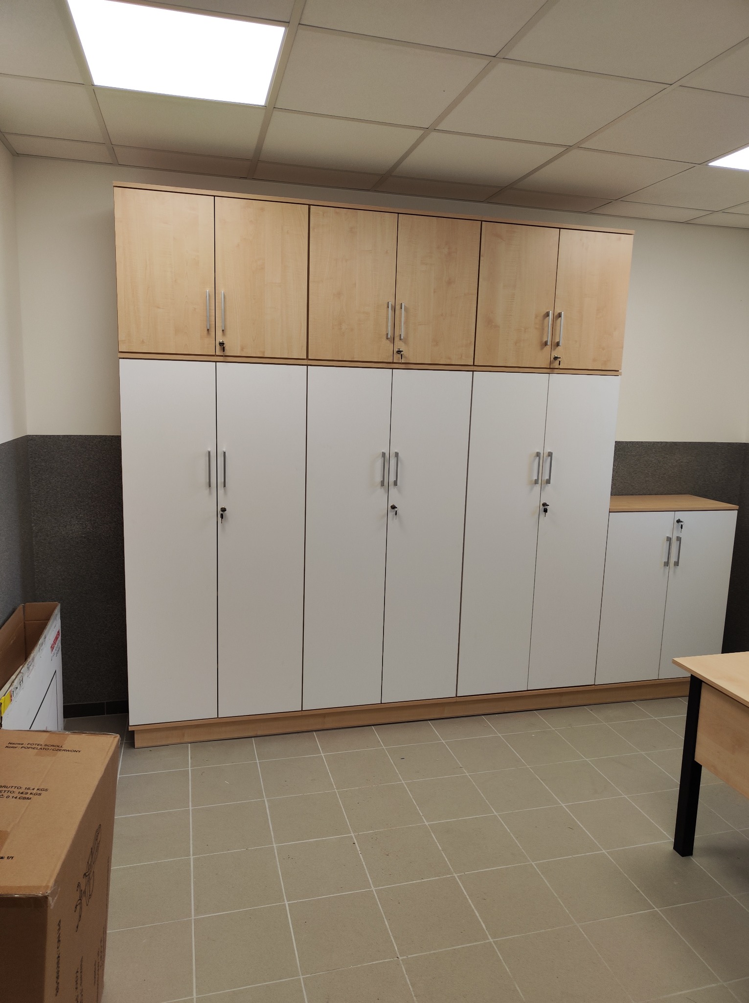 Wysokie szafy zamykane na klucz, białe dolne moduły i drewnopodobne górne, ustawione w rzędzie wzdłuż ściany, widoczne z boku biurko i karton z boku.