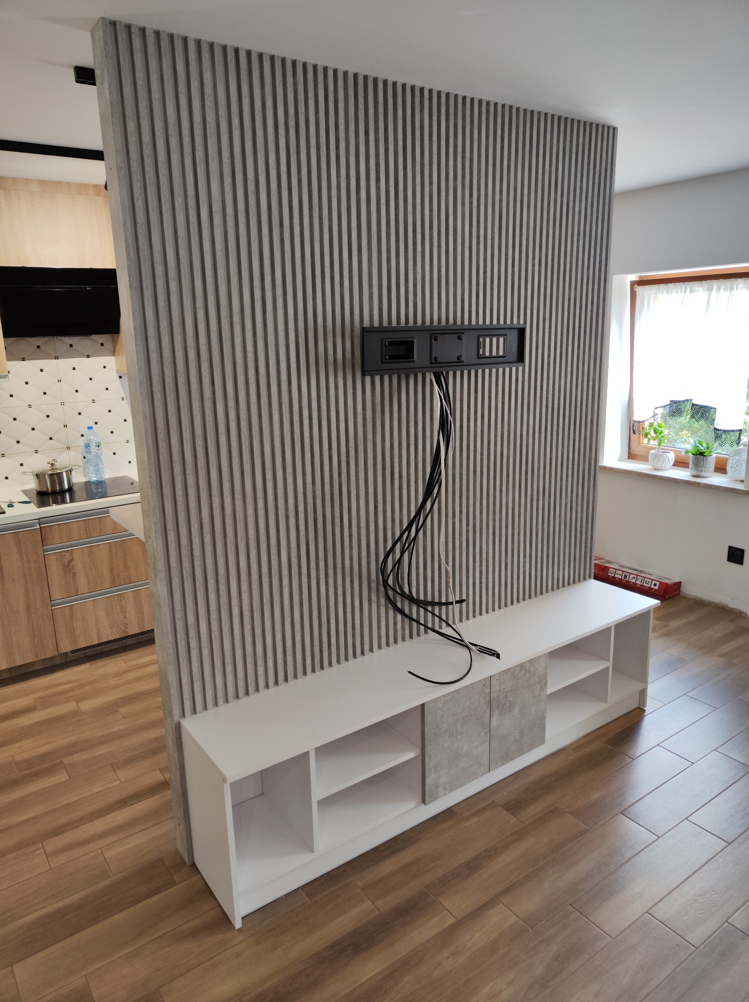 Nowoczesny salon z drewnianą podłogą, ścianą z paneli pionowych i białą szafką RTV z szarymi elementami, widoczne kable od telewizora i fragment kuchni w tle.