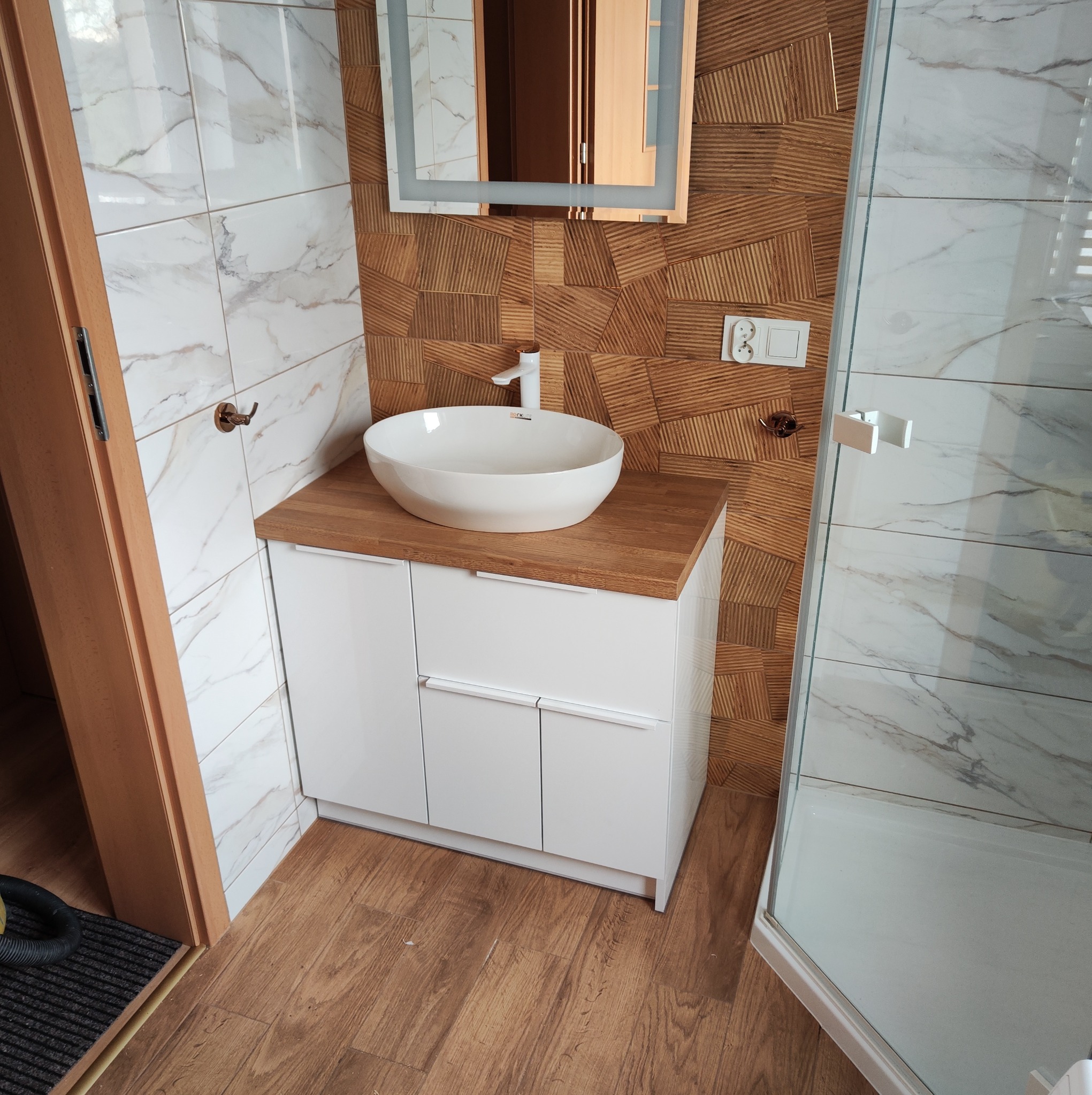 Nowoczesna łazienka z białą szafką pod umywalką nablatową na blacie z drewna, lustro z podświetleniem, ściany wyłożone płytkami imitującymi marmur i drewno, kabina prysznicowa w rogu pomieszczenia.