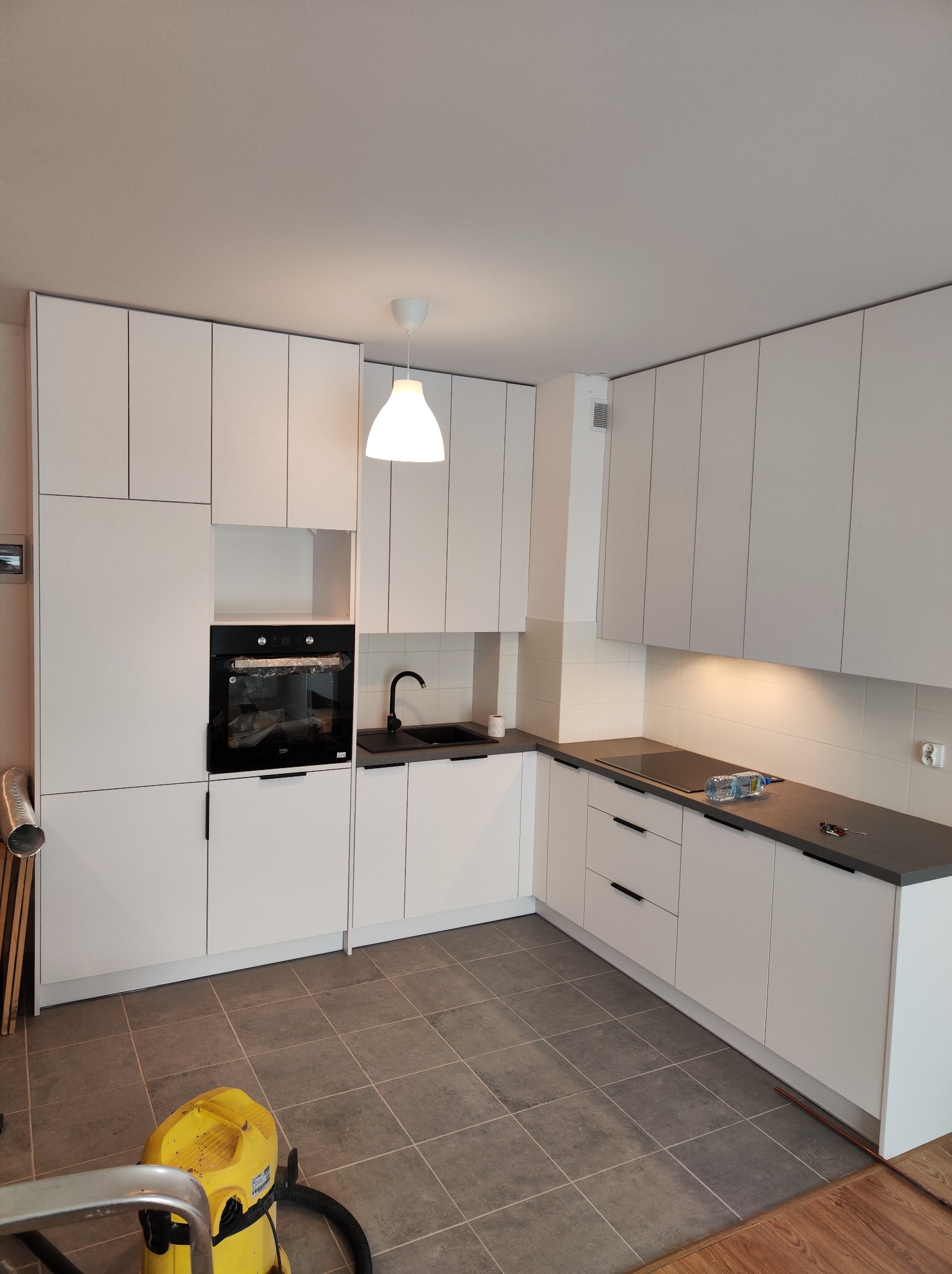 Nowoczesna, biała kuchnia z czarnymi akcentami po montażu, widoczny odkurzacz i drabina w tle, płytki podłogowe w szarym kolorze.