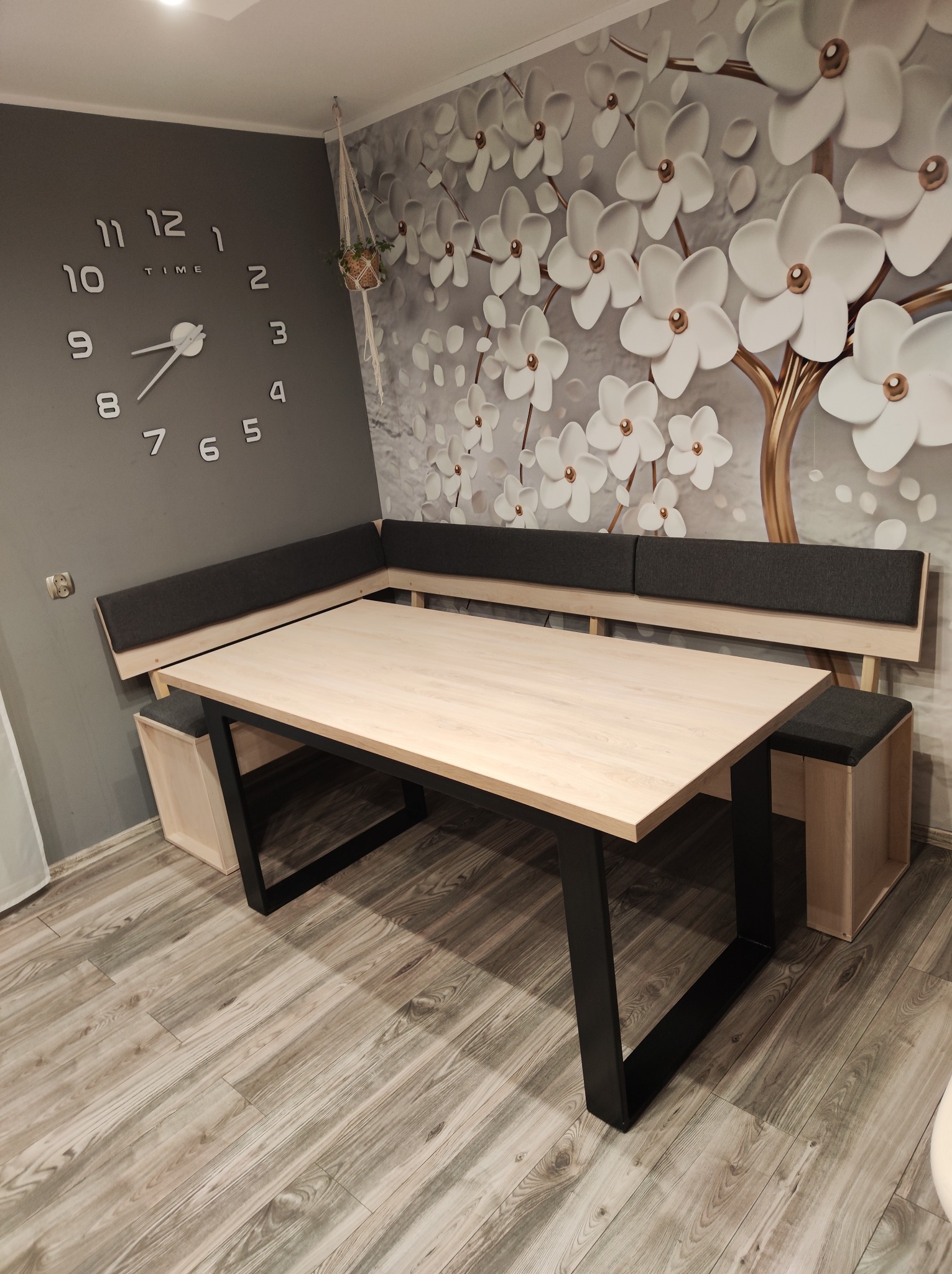 Drewniany stół z czarnymi metalowymi nogami i narożna ławka z szarym tapicerowanym oparciem oraz dwa taborety w jasnej kuchni z zegarem na ścianie i tapetą w kwiaty.