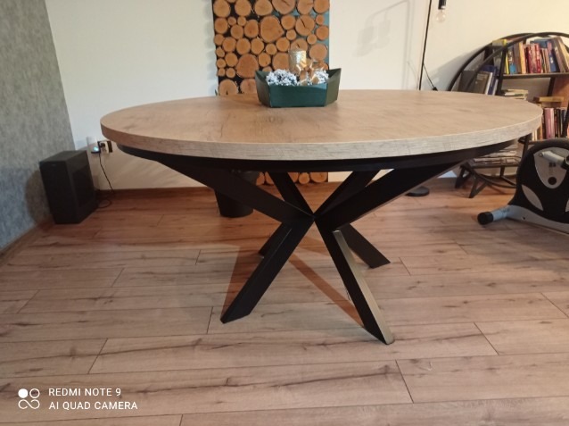 Okrągły stół z blatem w kolorze jasnego drewna i czarną, geometryczną podstawą, na drewnianej podłodze, z dekoracją na blacie i ścianą z drewnianych krążków w tle.