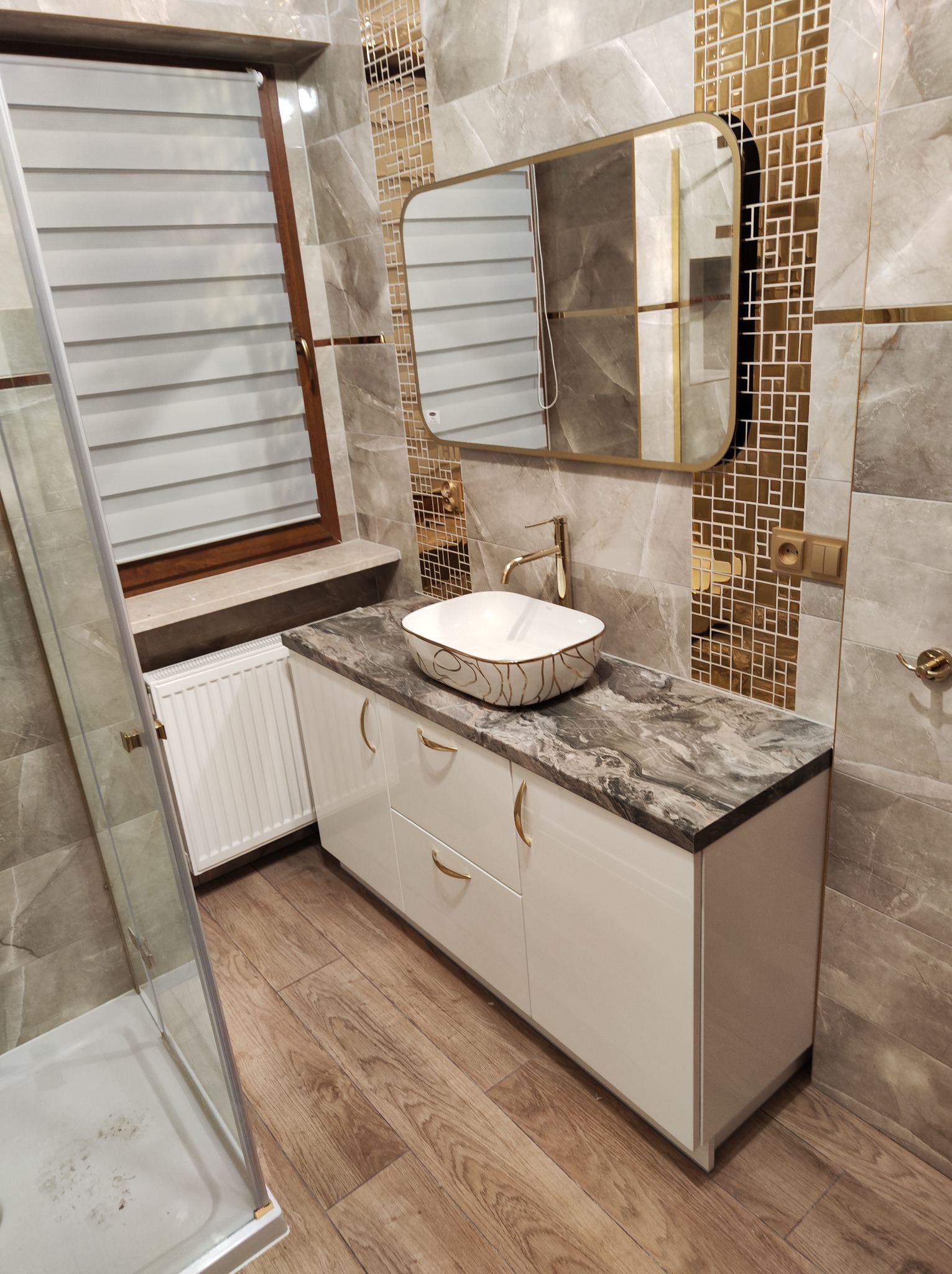 Nowoczesna łazienka z białą szafką pod umywalkę z marmurowym blatem, złotymi uchwytami i armaturą, lustrem w złotej ramie oraz dekoracyjnymi płytkami mozaikowymi w odcieniach złota i szarości.