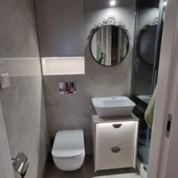 Nowoczesna łazienka z podświetlaną szafką pod umywalką, wiszącą toaletą i lustrem z ozdobnym obramowaniem. Szara płytka na ścianach i podłodze, kabina prysznicowa w tle.