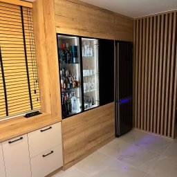 Ewel-Meb Ewelina Jasionowska Meble na wymiar - Nowoczesna zabudowa meblowa z barkiem i lodówką w salonie. Jasne drewno i beżowe fronty szafek tworzą spójną całość z drewnianymi lamelami na ścianie.