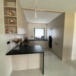 Jasna kuchnia z wyspą i otwartymi półkami, na których stoją dekoracje. Czarny blat kontrastuje z beżowymi frontami szafek i błyszczącą podłogą. Widoczne oświetlenie w stylu industrialnym.