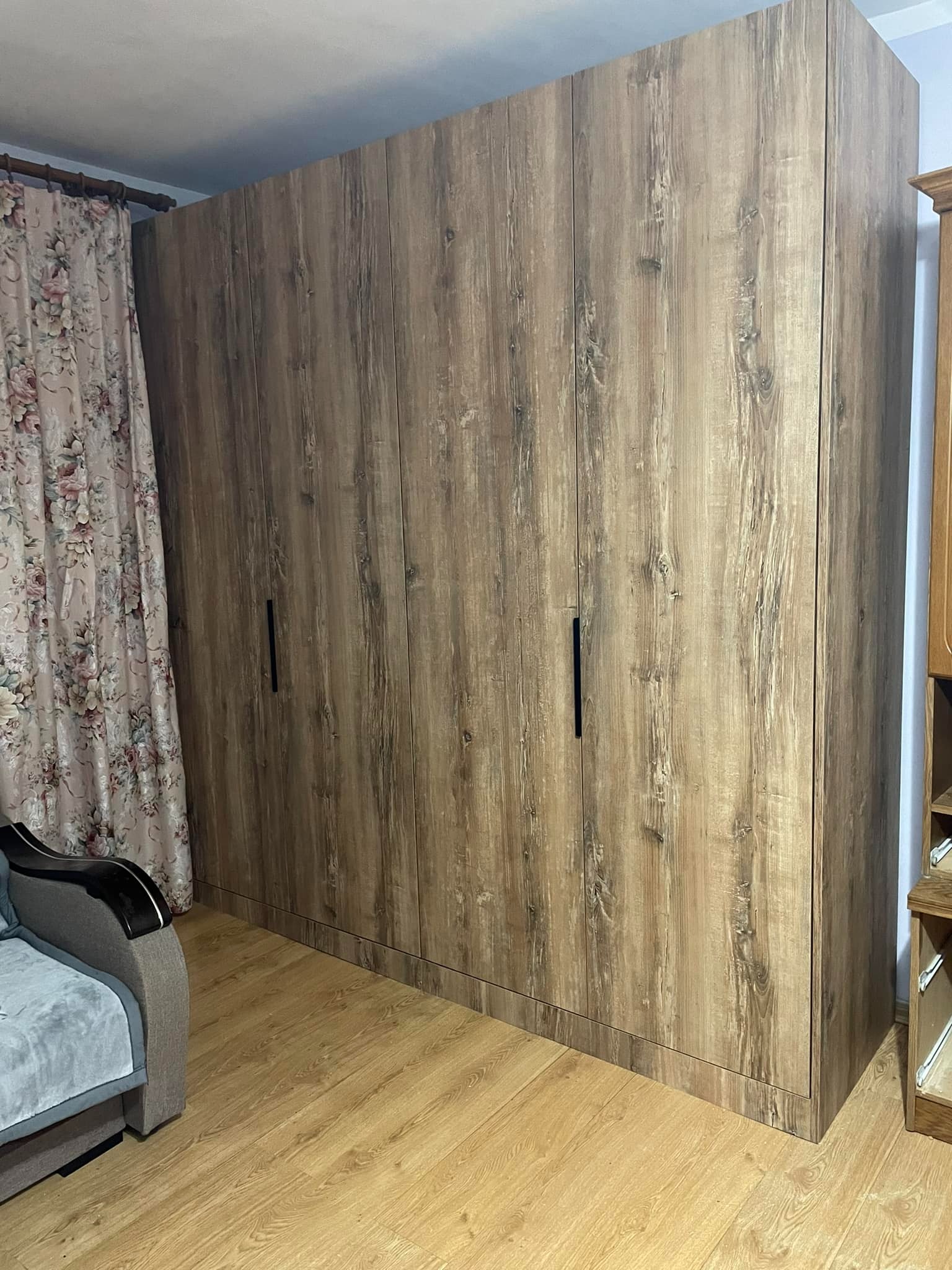 Wysoka szafa z drewnopodobnymi frontami i minimalistycznymi, czarnymi uchwytami, dopasowana do podłogi w kolorze drewna. Obok widoczny fragment sofy i zasłony.