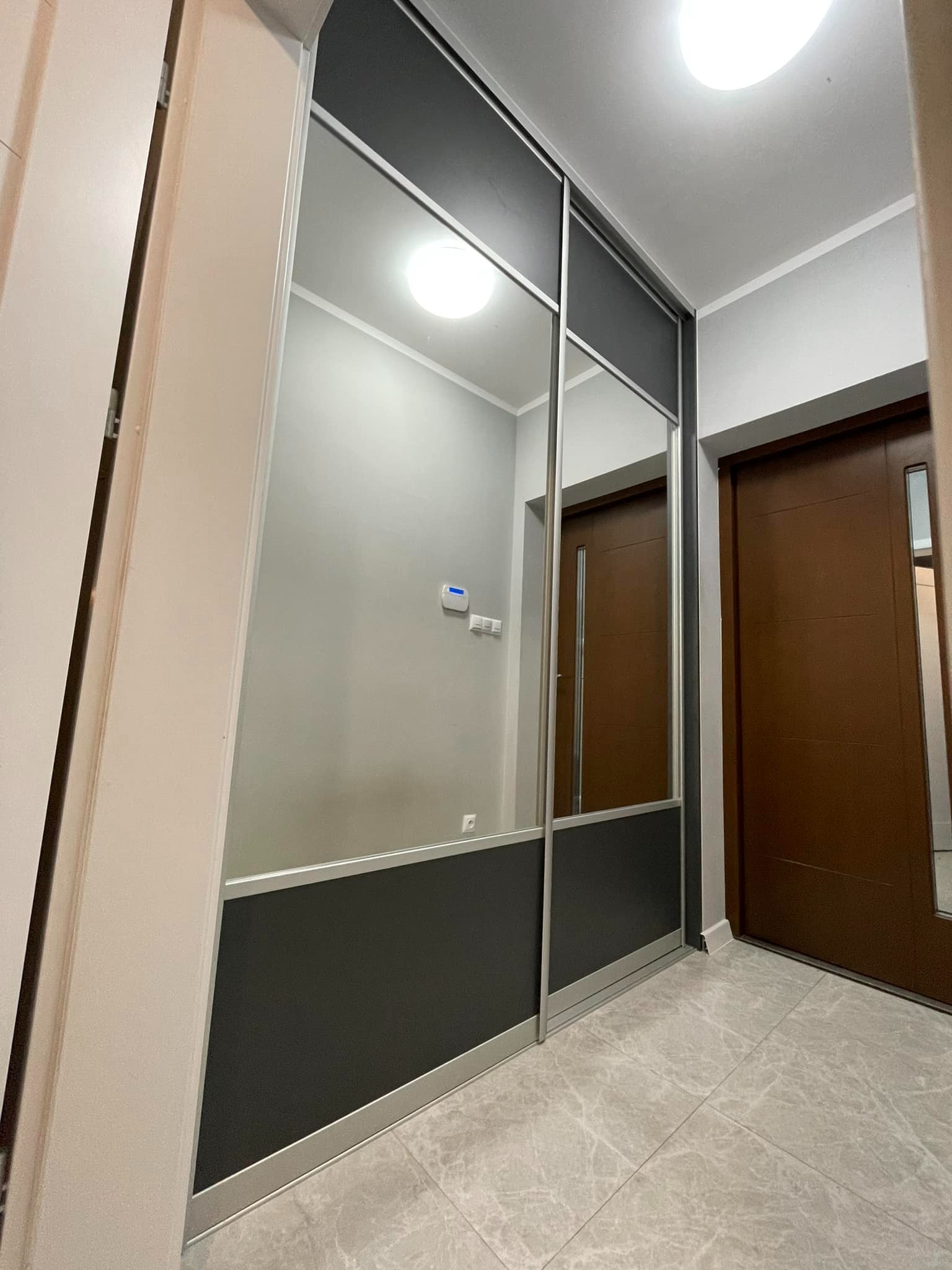 Nowoczesna szafa wnękowa z przesuwnymi, lustrzanymi drzwiami w korytarzu. Widoczne szare panele dolne i górne, odbicie drzwi wejściowych w lustrze szafy.