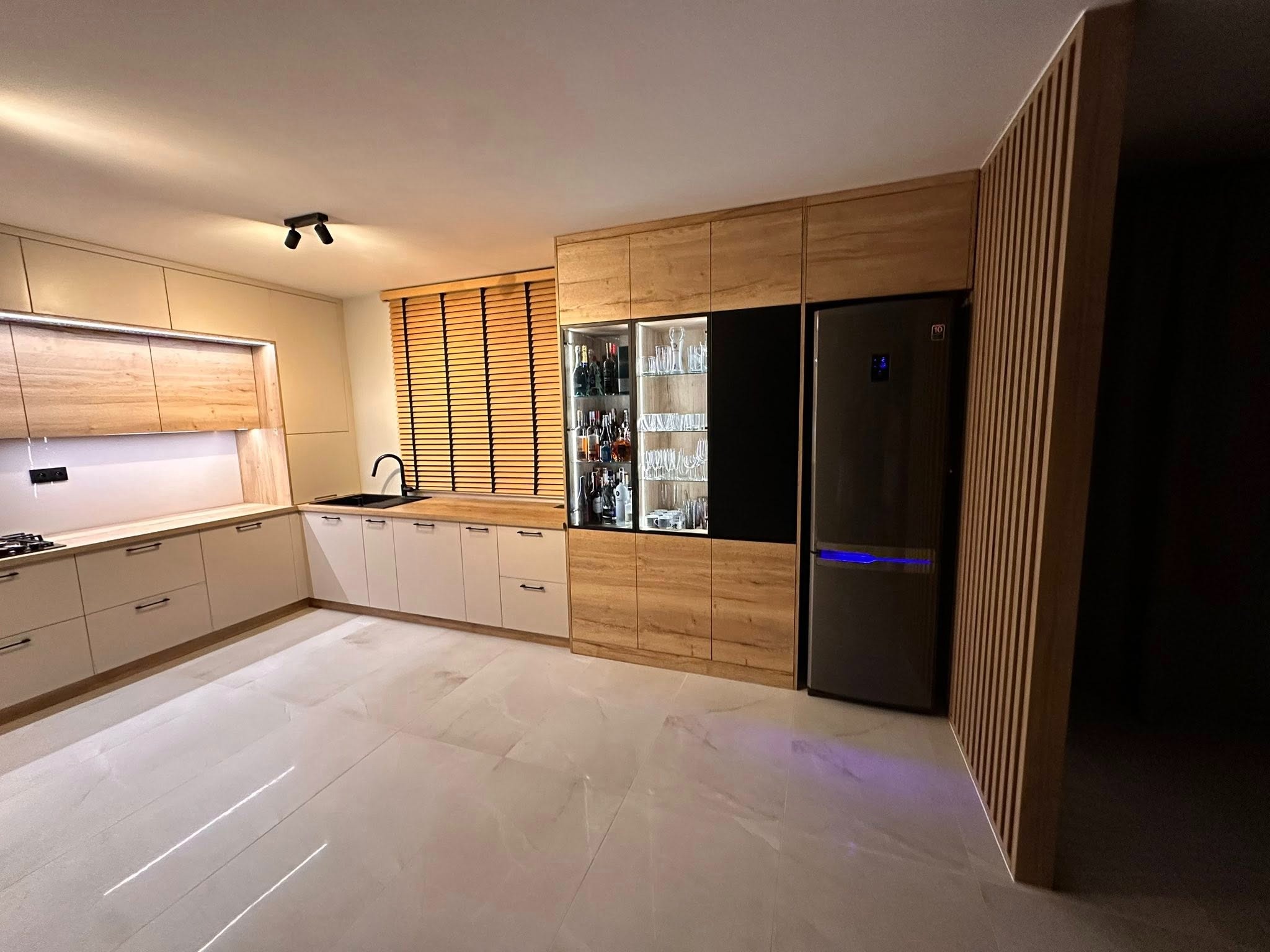 Nowoczesna kuchnia na wymiar z drewnianymi akcentami, zabudową lodówki i barkiem z oświetleniem, uchwyty listwowe, marmurowe płytki podłogowe.