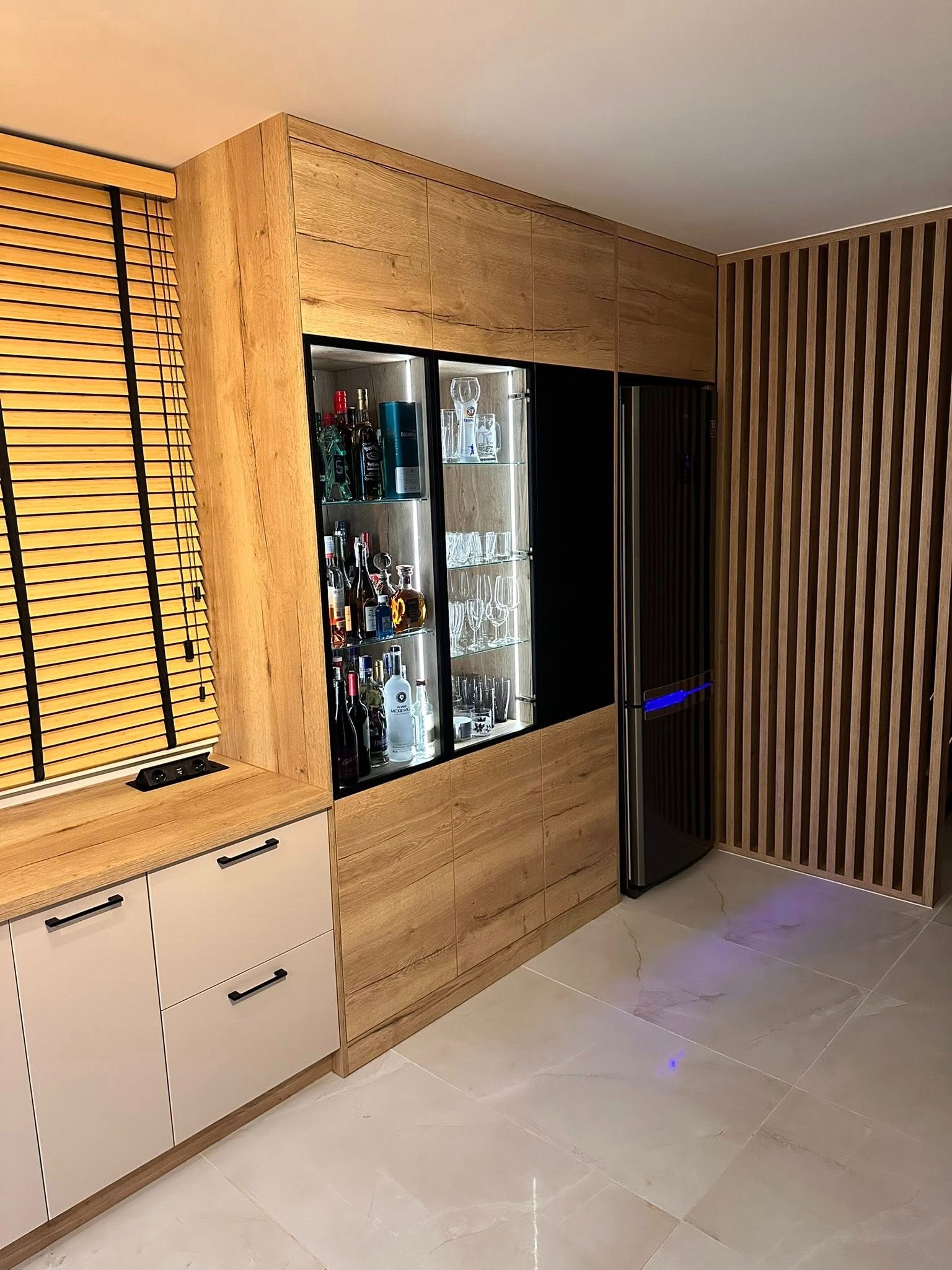 Nowoczesna zabudowa meblowa z barkiem i lodówką w salonie. Jasne drewno i beżowe fronty szafek tworzą spójną całość z drewnianymi lamelami na ścianie.