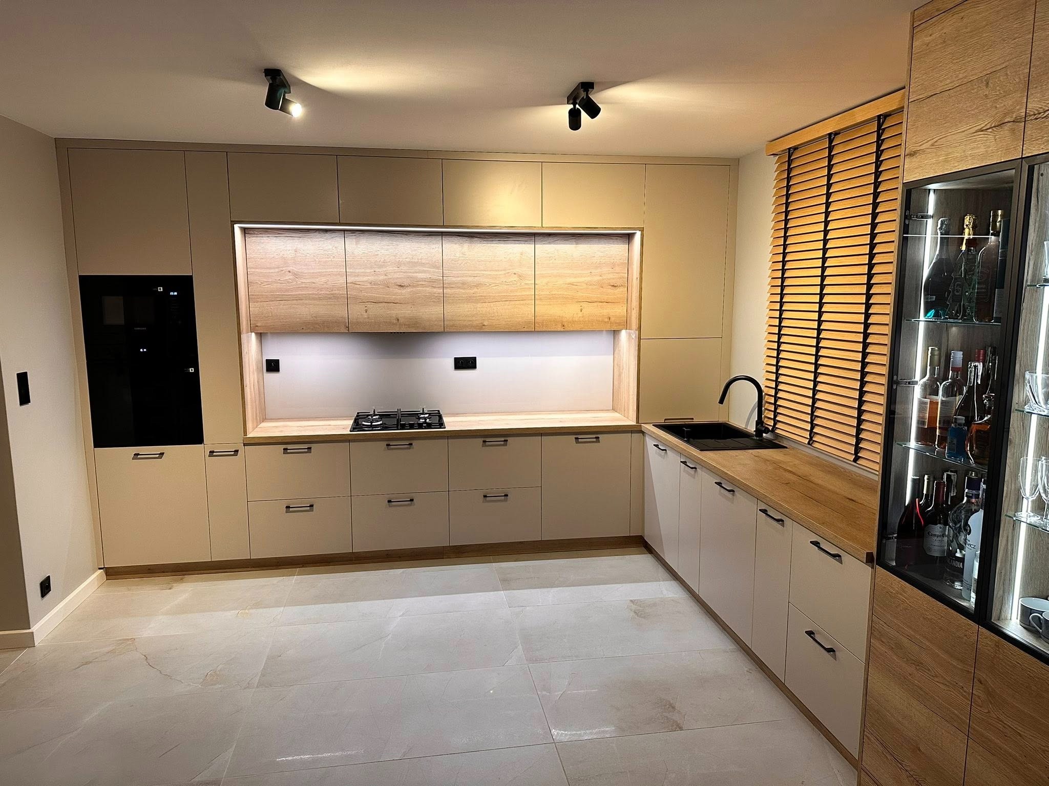 Jasna kuchnia na wymiar z drewnianym blatem i żaluzjami, oświetlenie punktowe. Fronty szafek w odcieniach beżu i kremu, nowoczesny design.