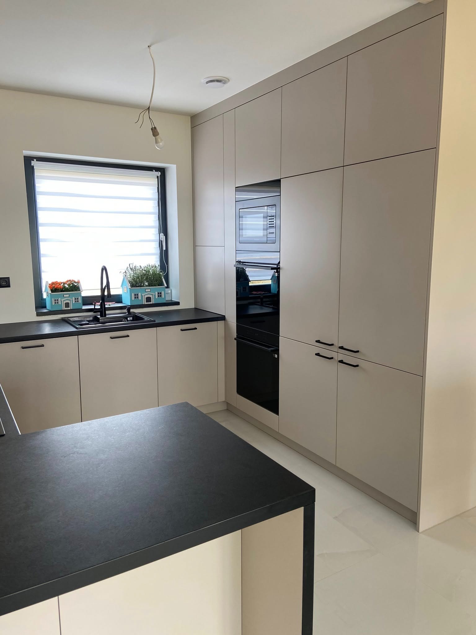 Jasna kuchnia z zabudową na wymiar, czarny blat i armatura. W oknie dekoracje w kształcie domków. Minimalistyczna lampa zwisa z sufitu.