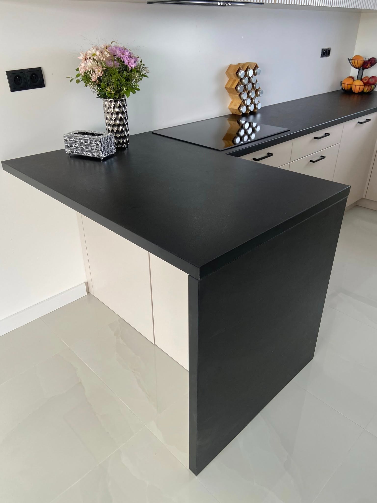 Nowoczesna kuchnia z czarnym blatem, jasnymi szafkami i dekoracyjnymi akcentami, takimi jak wazon z kwiatami i przyprawnik.