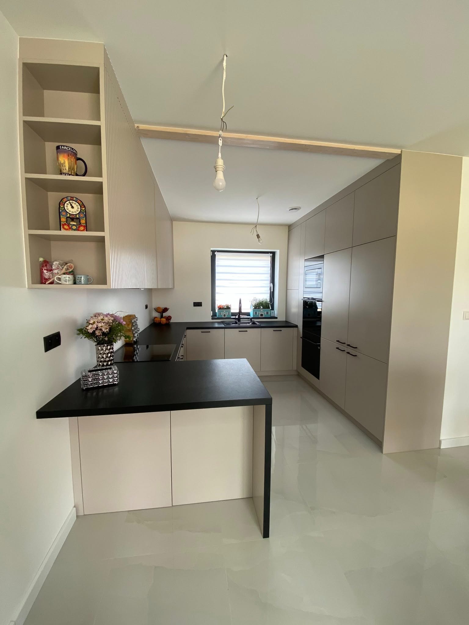 Jasna kuchnia z wyspą i otwartymi półkami, na których stoją dekoracje. Czarny blat kontrastuje z beżowymi frontami szafek i błyszczącą podłogą. Widoczne oświetlenie w stylu industrialnym.