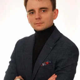 Portret młodego mężczyzny w granatowej marynarce i czarnym golfie, z zegarkiem na ręce i poszetką w kieszonce, na białym tle.