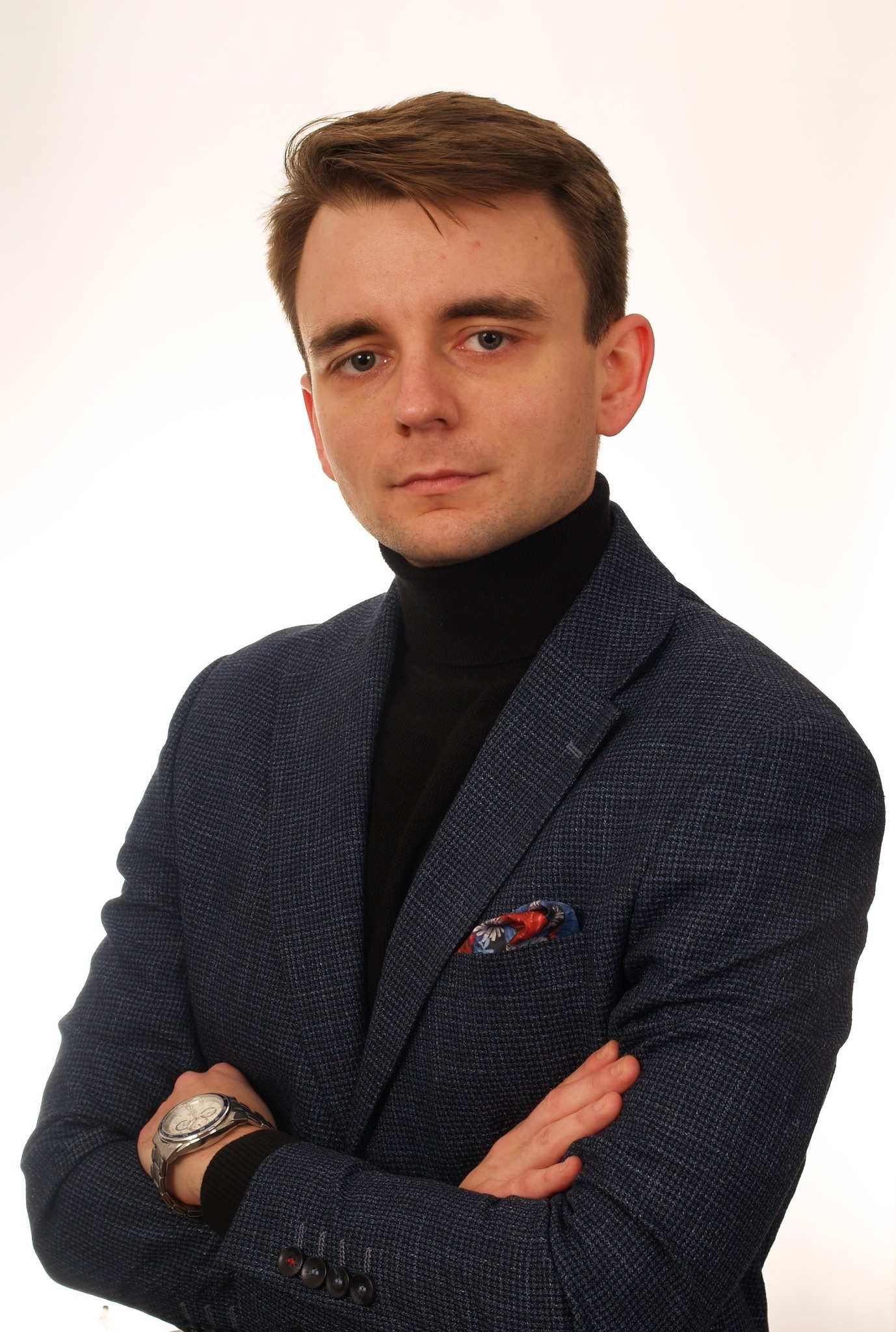 Portret młodego mężczyzny w granatowej marynarce i czarnym golfie, z zegarkiem na ręce i poszetką w kieszonce, na białym tle.