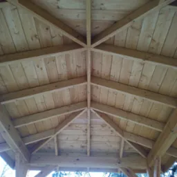 Wnętrze drewnianej altany ogrodowej z widoczną konstrukcją dachu, wykonaną z jasnego drewna, z deskami ułożonymi w jodełkę.