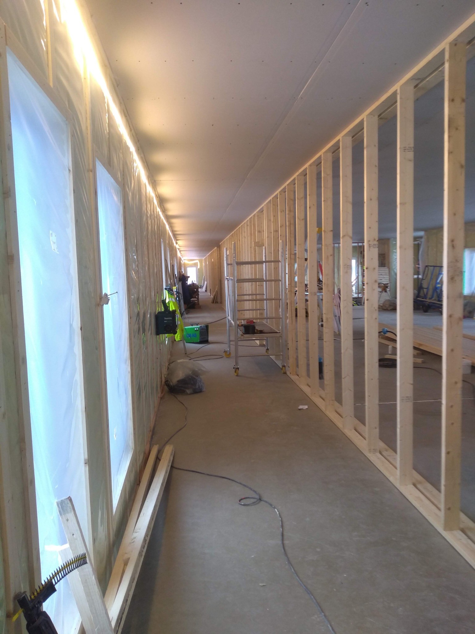 Wnętrze w trakcie budowy: widoczne drewniane ramy ścian działowych, okna zabezpieczone folią, narzędzia budowlane i drabina w tle.