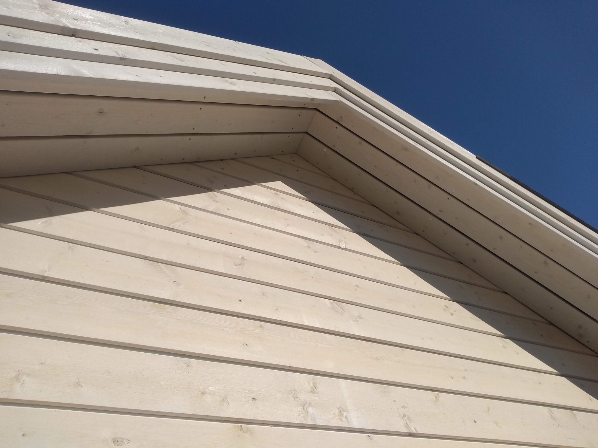 Fragment ściany zewnętrznej domu wykończonej jasnymi, drewnianymi deskami na pióro i wpust, widok z dołu pod kątem, na tle błękitnego nieba.