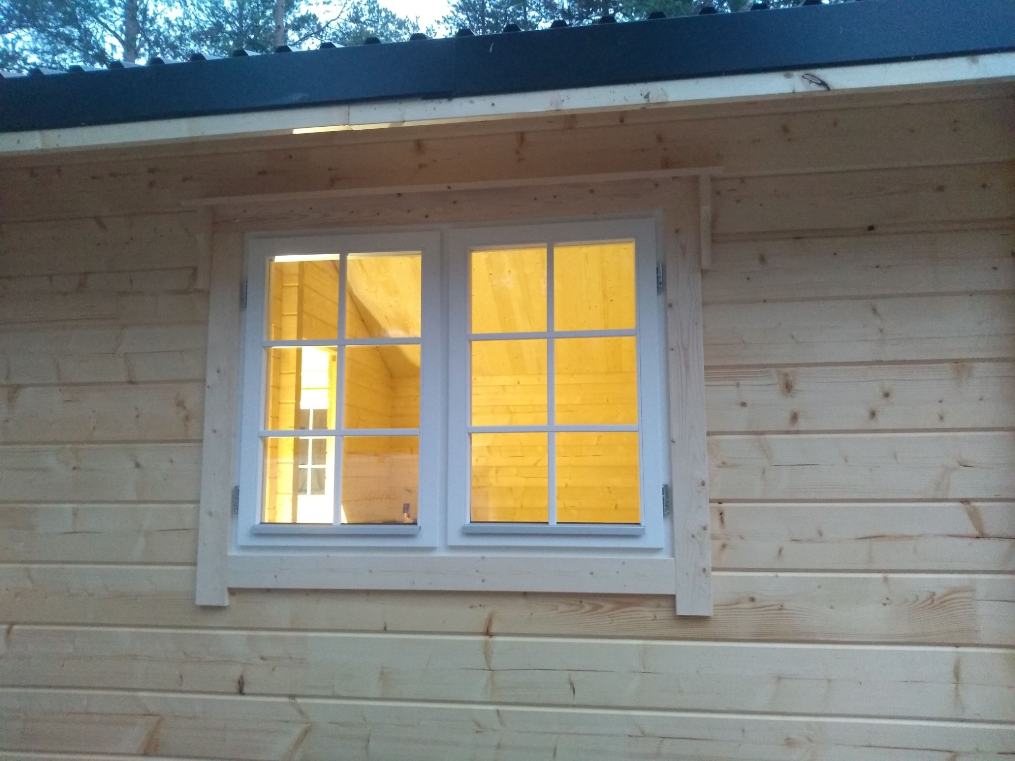 Fragment ściany zewnętrznej z jasnego drewna z oknem dwuskrzydłowym o białych ramach, przez które widać oświetlone wnętrze.