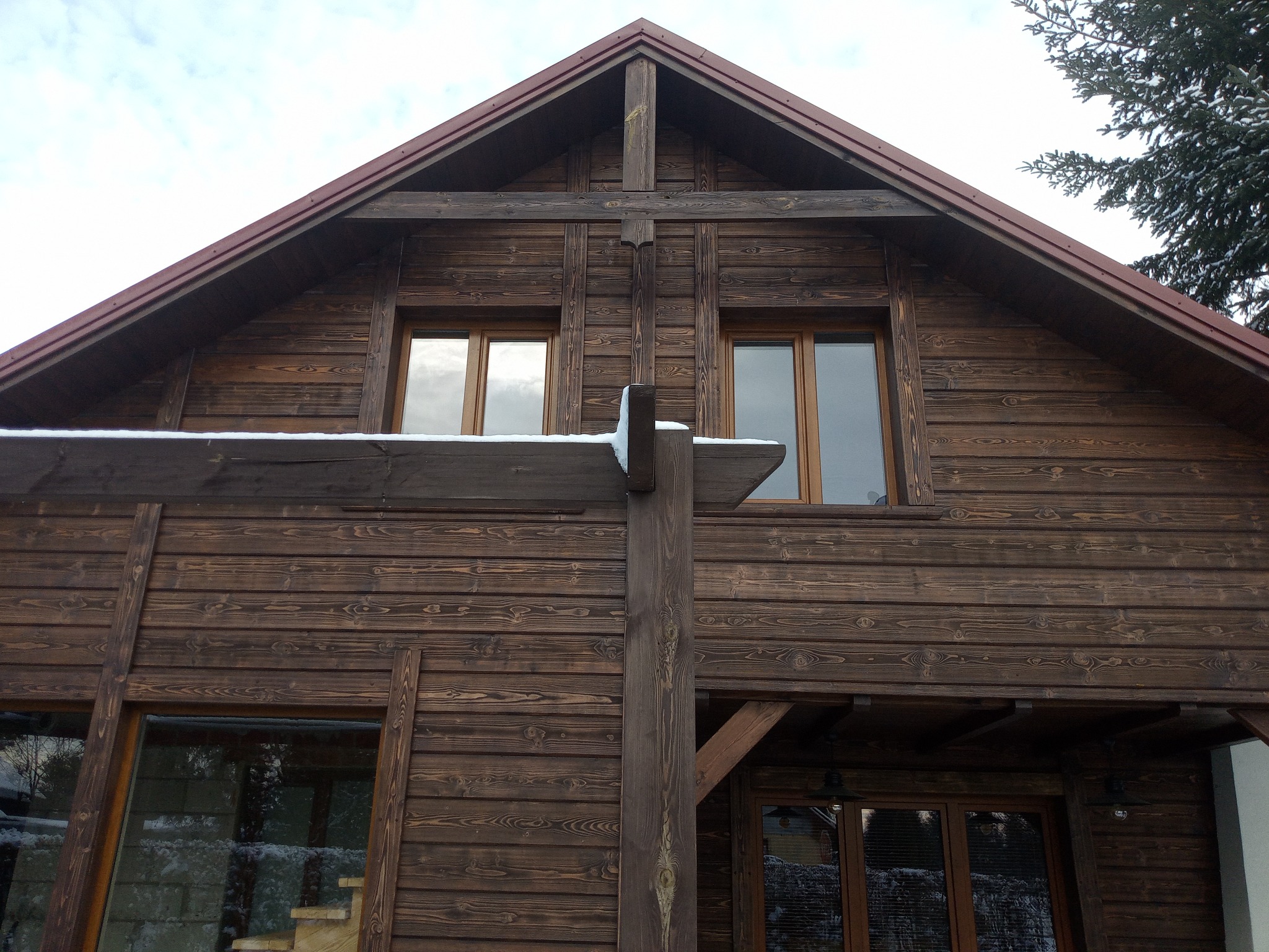 Fragment fasady domu z ciemnego drewna w Niedrzwicy Kościelnej z widocznymi elementami konstrukcyjnymi i oknami, częściowo pokryty śniegiem.
